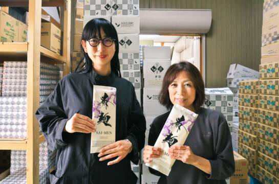 製麺所の製品の前で、「斎麺」を持つ南野マキさんと渡邉美千代さん。