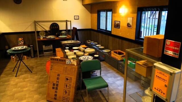 オレンジ色の照明の店内、中央に様々な大皿小皿が積まれたテーブル、奥にも模様入りの大皿が並んだ棚などがある。