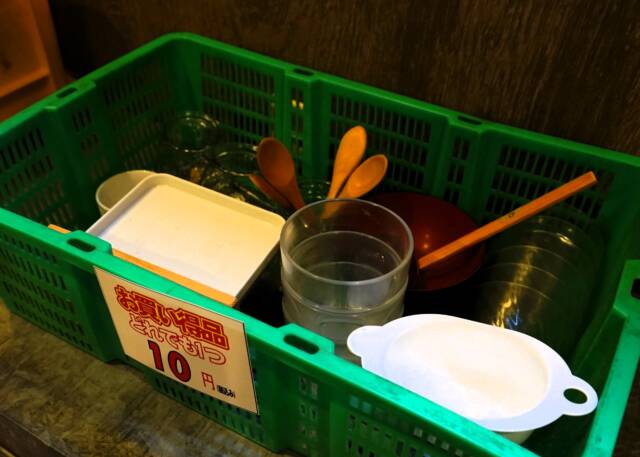 緑色の長方形のカゴに1つ10円のお買い得品のプラスチックのグラスや小さじ、小皿などが入っている