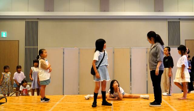 舞台上に6人、舞台袖には4人ほど子どもたちがいる。舞台の上では5人が立ち、中央では1人の少女が床にバレエダンサーのように開脚座りをして頬杖をついている。
