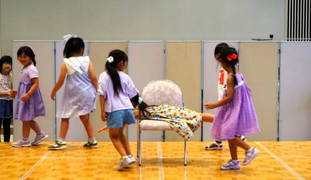 小さな女の子が7人、舞台上を走ったりしている。中央の椅子に1人が腹ばいで乗っている。