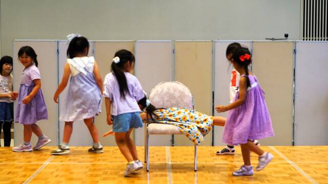 小さな女の子が7人、舞台上を走ったりしている。中央の椅子に1人が腹ばいで乗っている。
