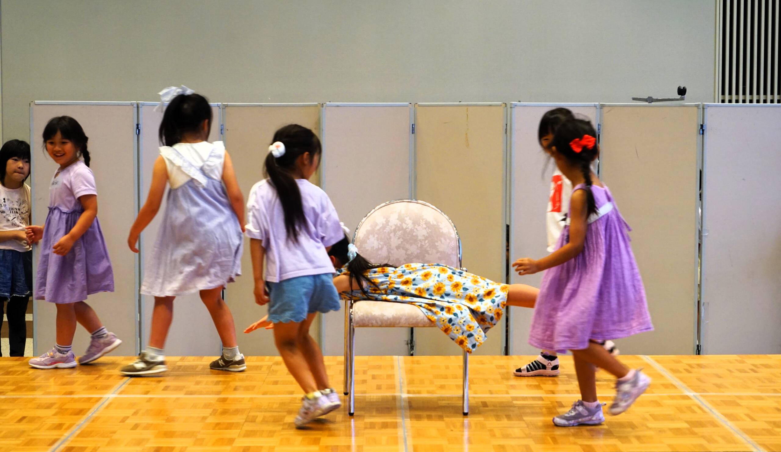 小さな女の子が7人、舞台上を走ったりしている。中央の椅子に1人が腹ばいで乗っている。