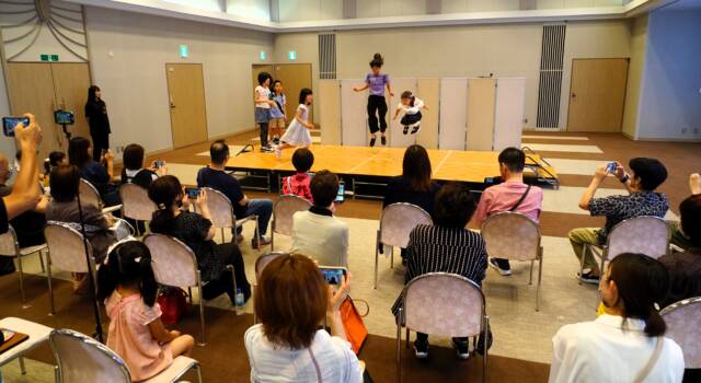 舞台上に5人の少女がいて、そのううちの2人がジャンプしている。観客席にはたくさんの人がいて、カメラを構えたりしている。