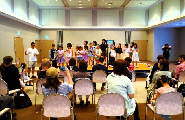 舞台上にたくさんの子どもたちが並んでいる。たくさんの観客は席について舞台を見ている。