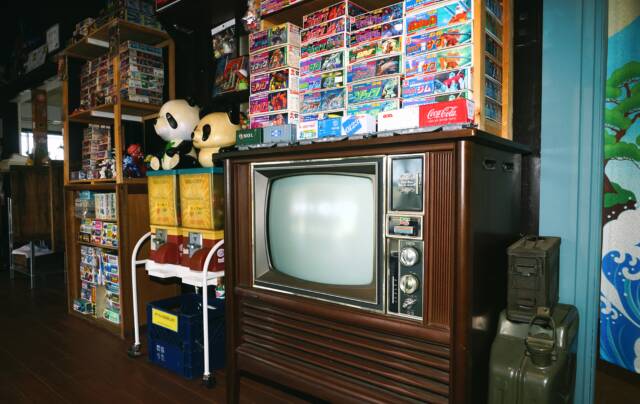 手前から大きな茶色い躯体のブラウン管テレビ、お手製の「昭和ガチャ」マシン、積み上げられたたくさんのプラモデルの箱など。