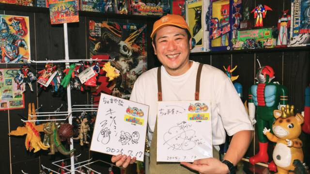 所狭しと並べられた昭和のおもちゃコレクションを前に、サイン色紙を両手に持ち笑顔の店主の姿