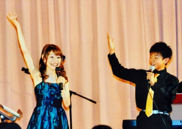 2013年の親子共演コンサートでの写真。右側に歌手の可愛アンリさん、左側に当時中学2年の舞台俳優・山本航大さんが写っている。二人は笑顔で歌い、左手にマイク、右手を挙げてポーズしている。