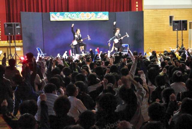 海蔵小学校の体育館で、児童と教員が一緒に手を高く掲げてライブを楽しむ様子。