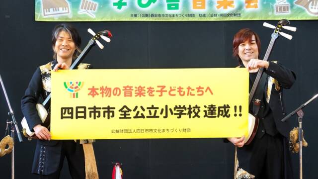 四日市市出身のKUNI-KENが、海蔵小学校の体育館ステージで「四日市市　全公立小学校達成」と書かれた横断幕を掲げている様子。