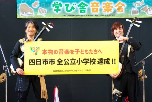 四日市市出身のKUNI-KENが、海蔵小学校の体育館ステージで「四日市市 全公立小学校達成」と書かれた横断幕を掲げている様子。