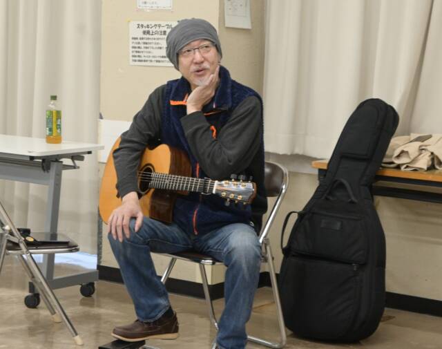 ギターを膝に乗せ手で顔を触れて話す竹内さん