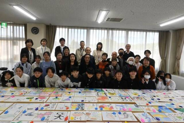 色紙の後に立つ民生委員・児童委員と三滝中学校富平中学校の生徒と神前小学校の児童の記念写真