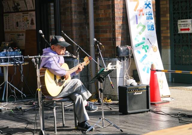 ギターを手に歌うEIJIさん。ウッドデッキのステージで観客に向けて歌唱している