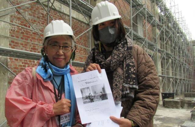 ヘルメットをかぶった出口さんと台湾人通訳の女性が工場跡地にYOUよっかいちの記事を手に立っている