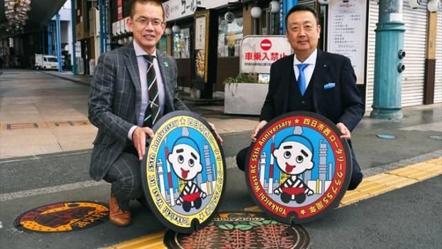 諏訪栄町の歩道でデザインマンホールのレプリカを持つ佐野貴信会長と田中大補実行委員長