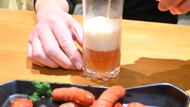 ウインナーが盛り付けられた皿の後ろでクラフトビール「琥珀のとき」をグラスに注いでいる