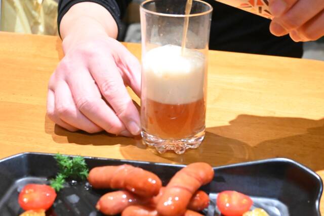 ウインナーが盛り付けられた皿の後ろでクラフトビール「琥珀のとき」をグラスに注いでいる