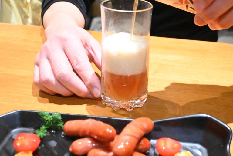 ウインナーが盛り付けられた皿の後ろでクラフトビール「琥珀のとき」をグラスに注いでいる