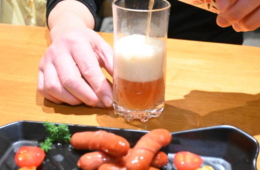 ウインナーが盛り付けられた皿の後ろでクラフトビール「琥珀のとき」をグラスに注いでいる