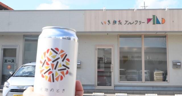 クラフトビール「琥珀のとき」を手にし、いきかたブルワリーの前に立つ人の手と缶