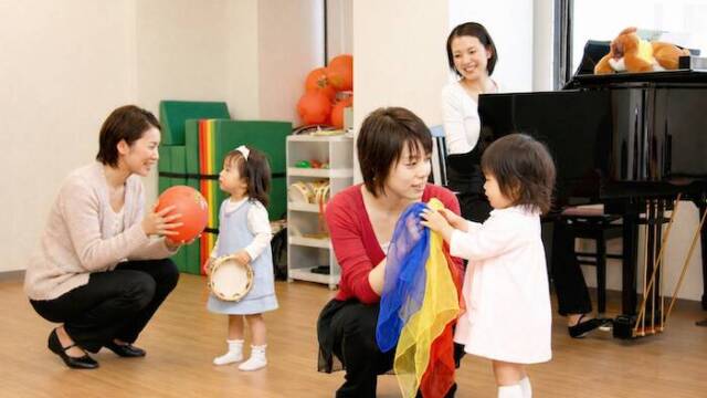 カワイ音楽教室の幼児レッスン。カラフルなスカーフ、ボール、タンバリンで遊ぶ親子と講師。奥にグランドピアノ。