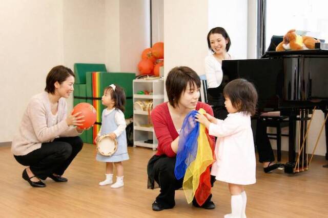 カワイ音楽教室の幼児レッスン。カラフルなスカーフ、ボール、タンバリンで遊ぶ親子と講師。奥にグランドピアノ。