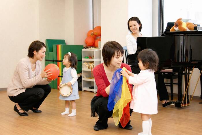 カワイ音楽教室の幼児レッスン。カラフルなスカーフ、ボール、タンバリンで遊ぶ親子と講師。奥にグランドピアノ。