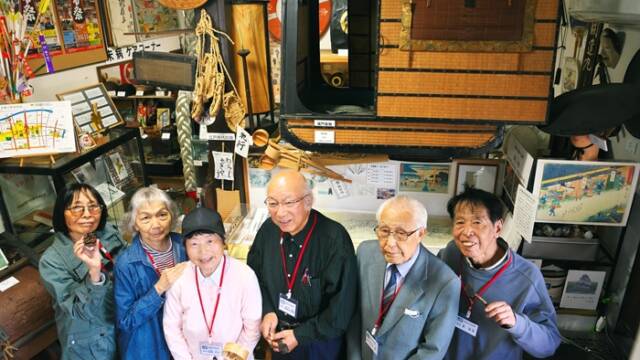 東海道四日市宿資料館で、江戸時代の駕籠の空中展示を背景に並ぶ長谷川博久館長と語り部ボランティアたち