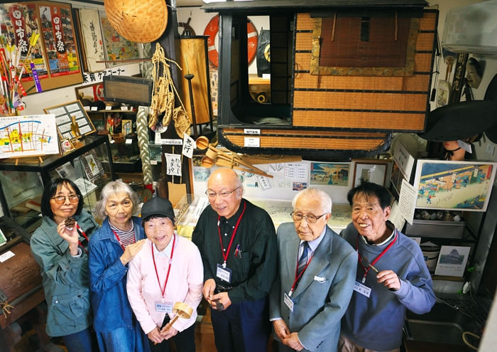 東海道四日市宿資料館で、江戸時代の駕籠の空中展示を背景に並ぶ長谷川博久館長と語り部ボランティアたち