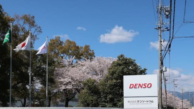 満開の桜と青空を背景にした株式会社デンソートリムの看板と工場外観。