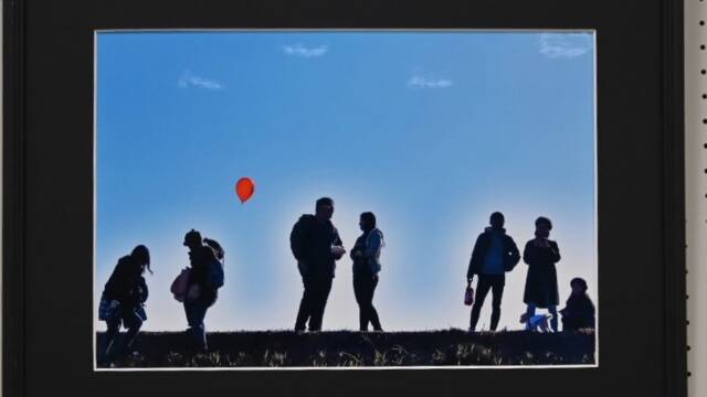 青空を背景に7人の人が立つ赤い風船が印象的な写真