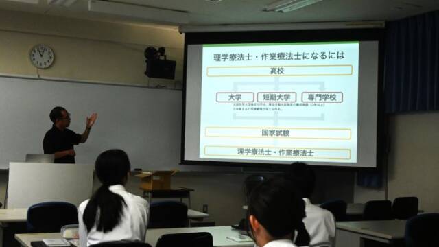 進路ガイダンスで理学療法士と作業療法士の説明が写ったスクリーンと話を聞く生徒