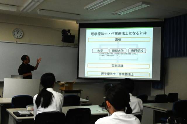 進路ガイダンスで理学療法士と作業療法士の説明が写ったスクリーンと話を聞く生徒