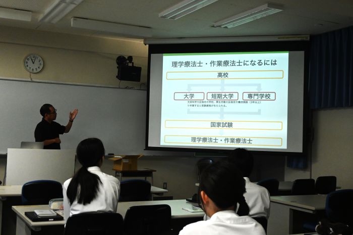 進路ガイダンスで理学療法士と作業療法士の説明が写ったスクリーンと話を聞く生徒