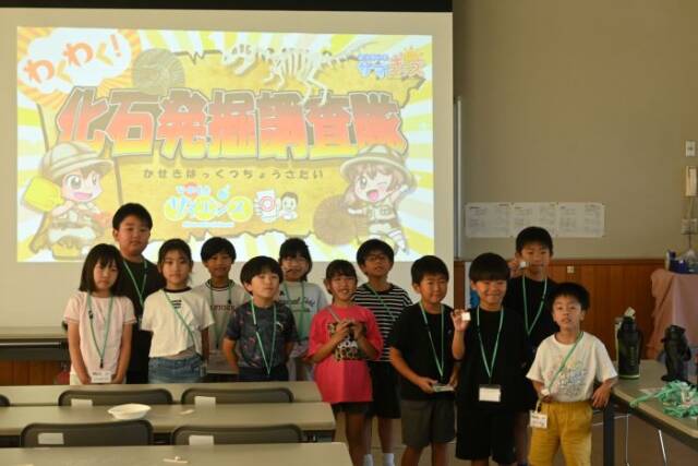 化石発掘調査隊と写し出されたスクリーンの前で記念写真を撮る子どもたち