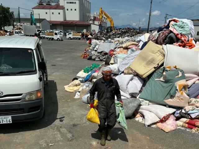 被災し使用不可能になった寝具がうず高く積まれた災害ゴミ集積場で両手に持ったゴミ袋を運ぶ石川さん