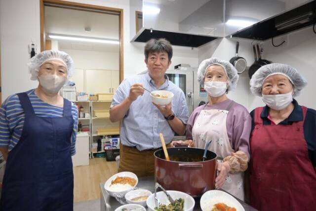 昼食づくりの高齢者ボランティアとカレーを持ちながら写真に納まる貝沼院長