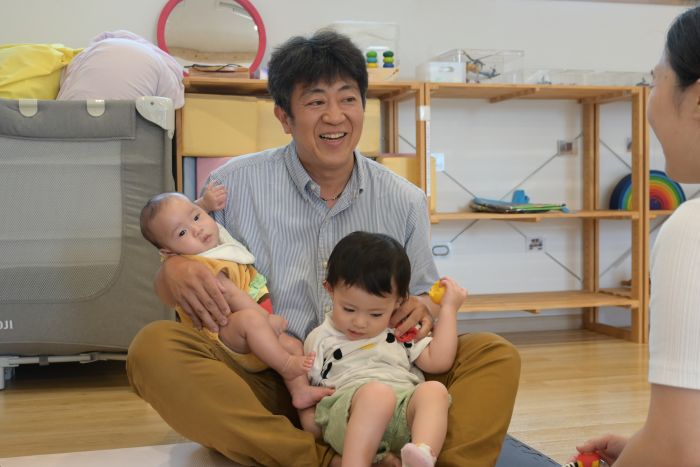 赤ちゃんを膝に乗せ片方の手で別の赤ちゃんを抱き、笑顔で母親と話す貝沼院長