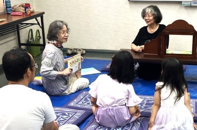 紙芝居コーナーで、女性2人が紙芝居を披露し、子ども2人と男性が向かい合って鑑賞している場面。