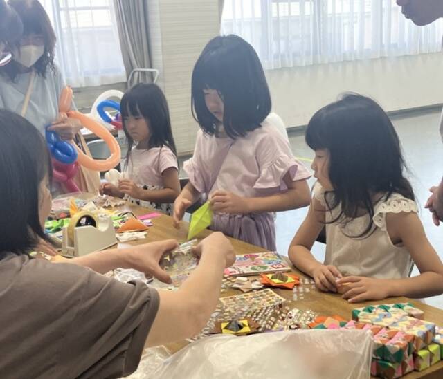 折り紙コーナーで、子どもと大人が一緒に色紙を折りながら交流している場面。