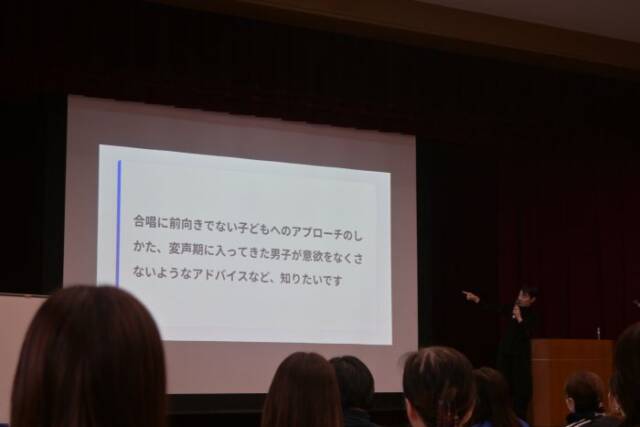 合唱に前向きでない子どもへのアプローチの仕方など質問が写りだされたスクリーンを指さす田中さん