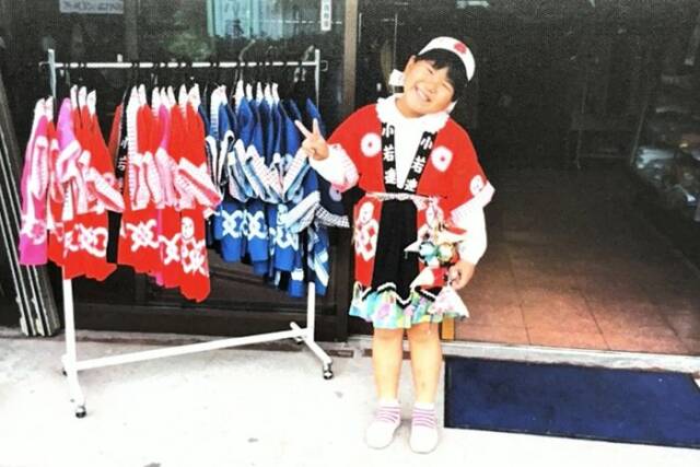 祭りの法被を着た笑顔の子どもが、店頭でピースサインをしている。横には法被が並んだハンガーラックがある。