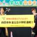四日市市出身のKUNI-KENが、海蔵小学校の体育館ステージで「四日市市　全公立小学校達成」と書かれた横断幕を掲げている様子。